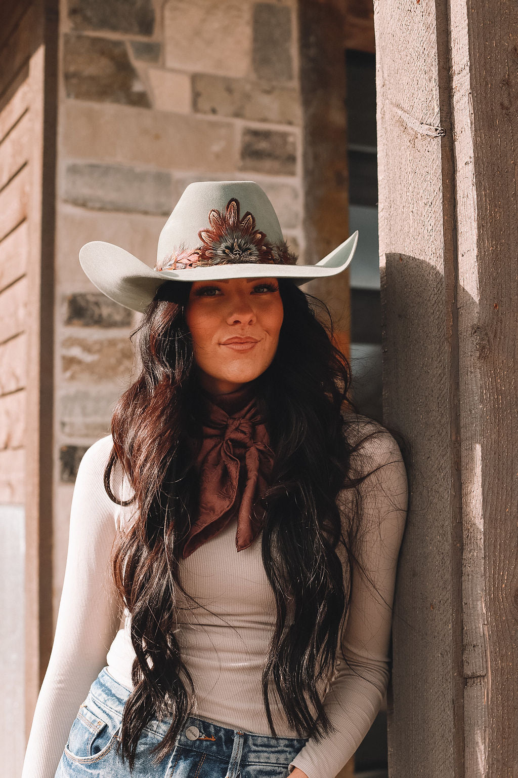 Woman in a teal cowgirl hat wearing a handmade feather hatband and medallion, styled with a wild rag and confident smile.