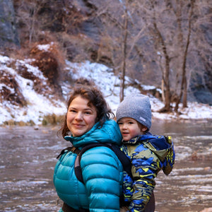 Woman and child in winter clothing standing by a snowy stream, at the entrance to the Narrows in Zion National Park, with trees and rocks in the background.