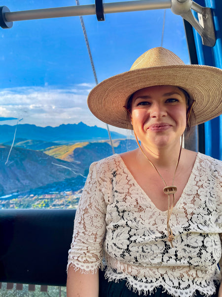 Olivia Poyer riding a gondola in Jackson Hole, wearing a lace blouse and straw hat, with the Teton mountain range in the background.