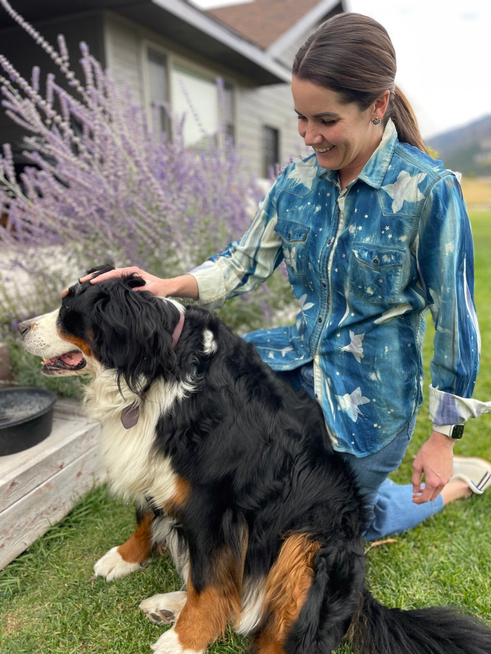 Woman in a whimsical floral cyanotype shirt petting her dog, smiling with purple flowers and mountains in the background.