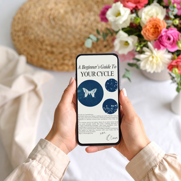 Hands holding a phone displaying Olivia Poyer’s free cycle tracking guide, styled on a neutral tablecloth with a bouquet of pink, orange, and white flowers.