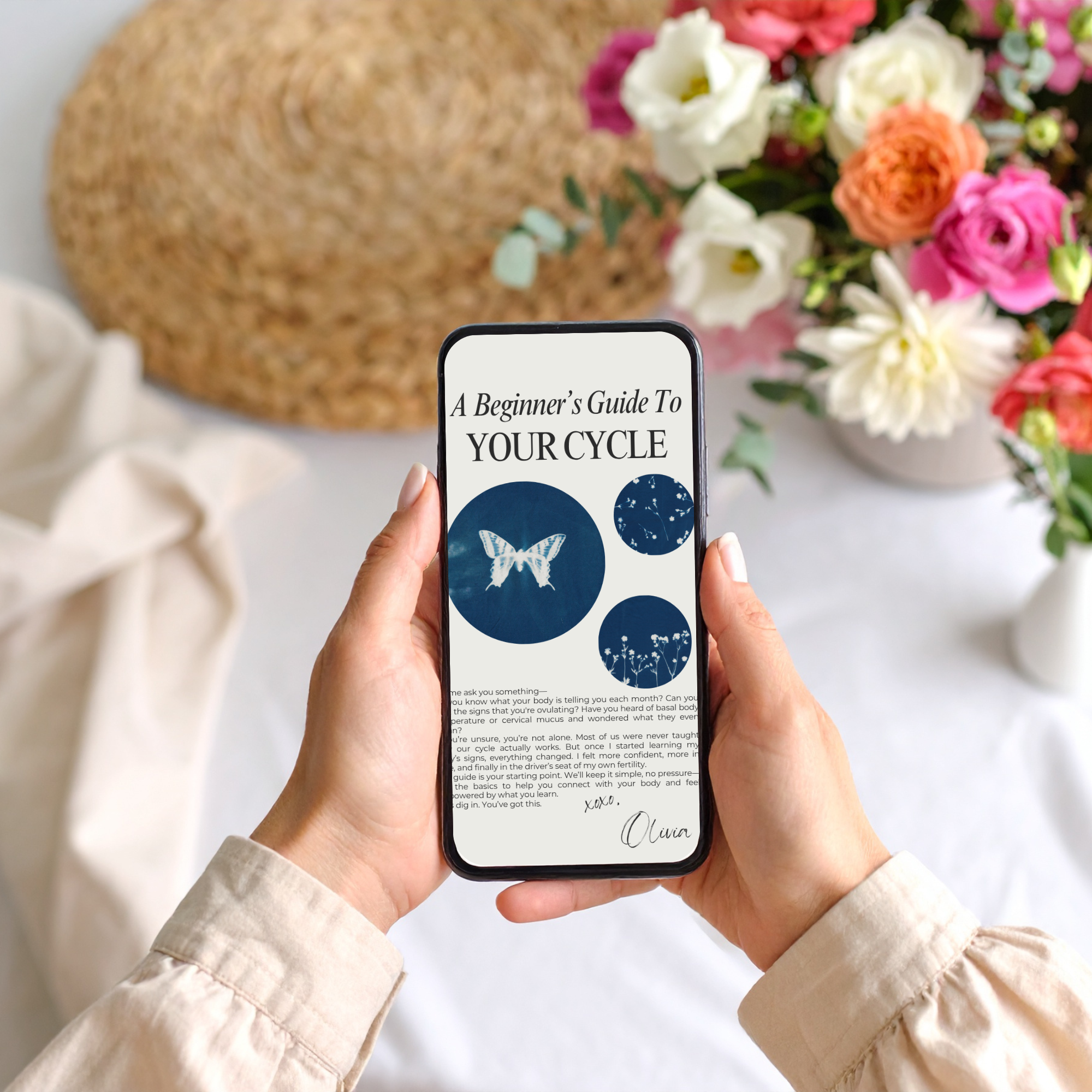 Hands holding a phone displaying “A Beginner’s Guide to Your Cycle,” with flowers and woven placemat in background — modern, natural lifestyle imagery.