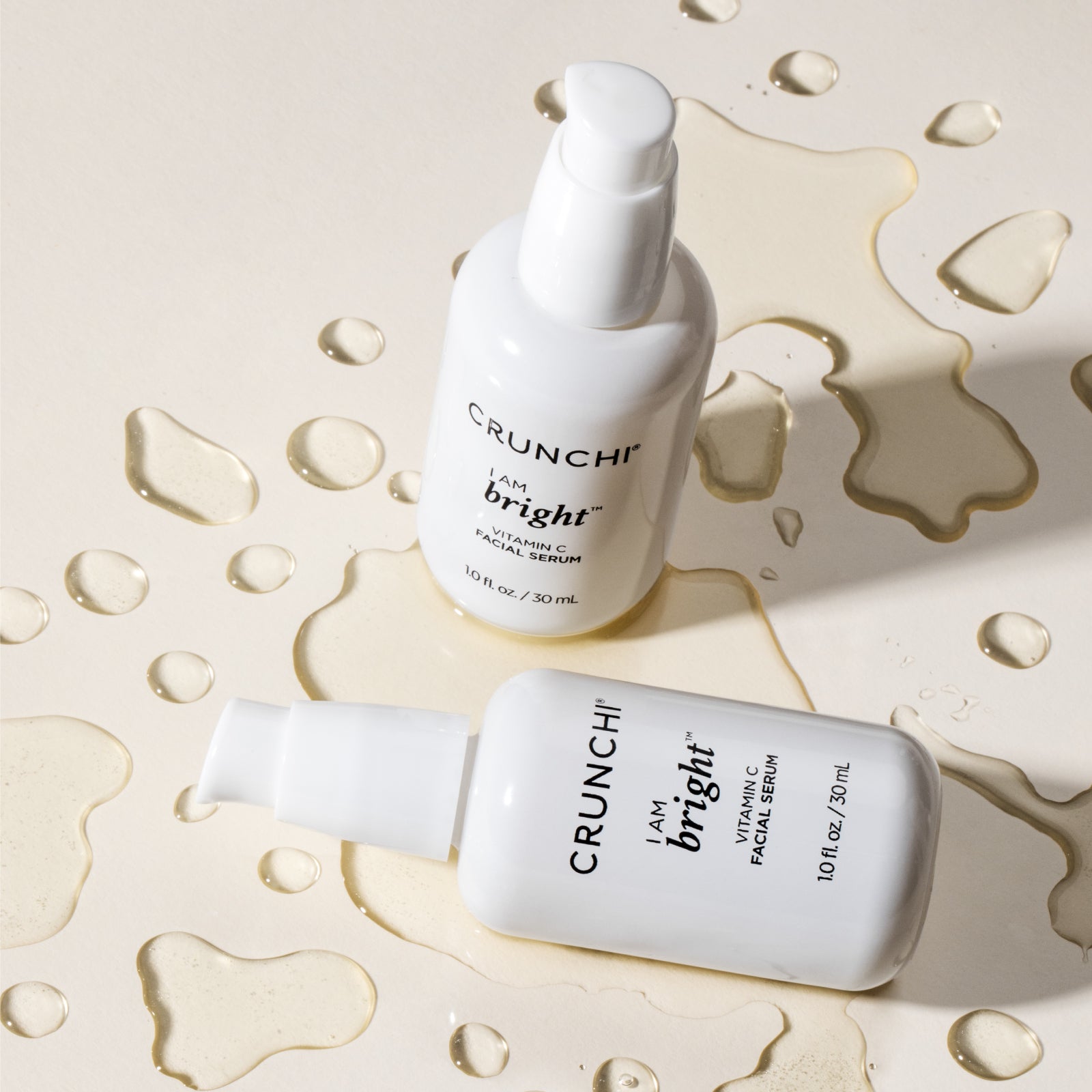 Two bottles of Crunchi I AM Bright Vitamin C Serum on a neutral backdrop, one tipped over with product pooling artistically around it.