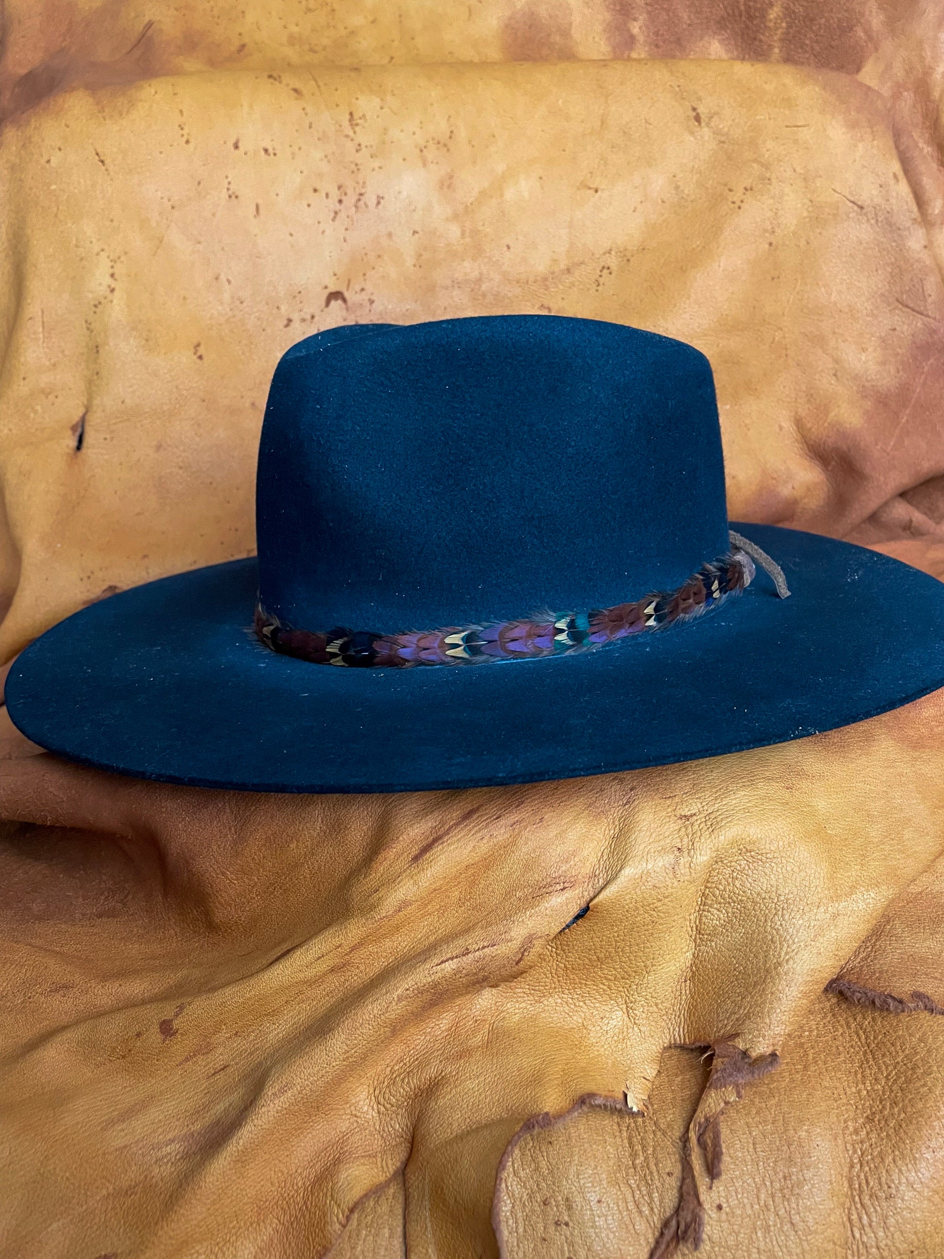 A black felt hat adorned by a pheasant feather hatband. The feathers are arranged in a repeating pattern: Maroon, Gold, Emerald. 