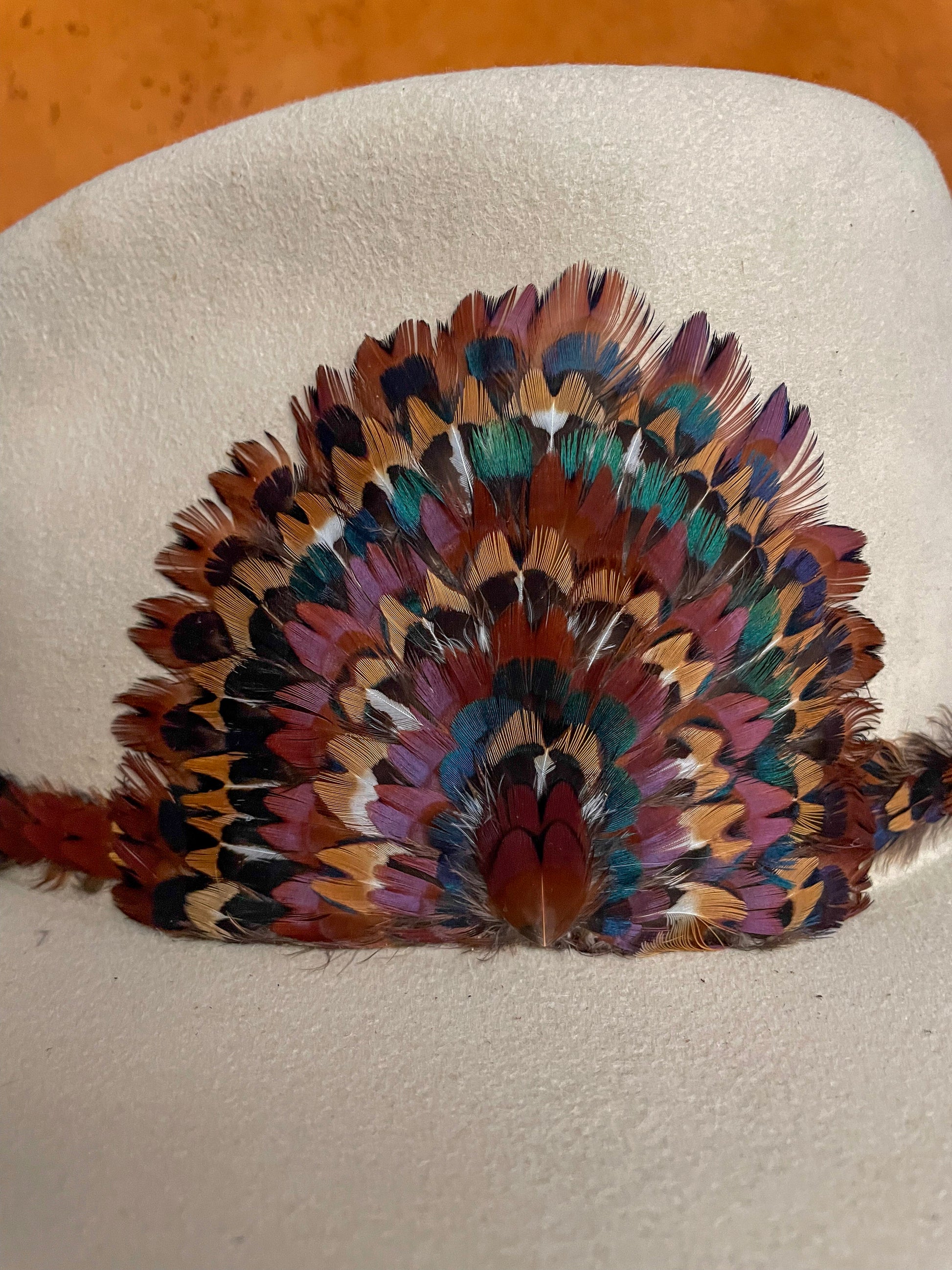 A close up side view of a handcrafted hatband and removable feather medallion built from the iridescent neck feathers of a wild rooster pheasant on an ivory felt hat.