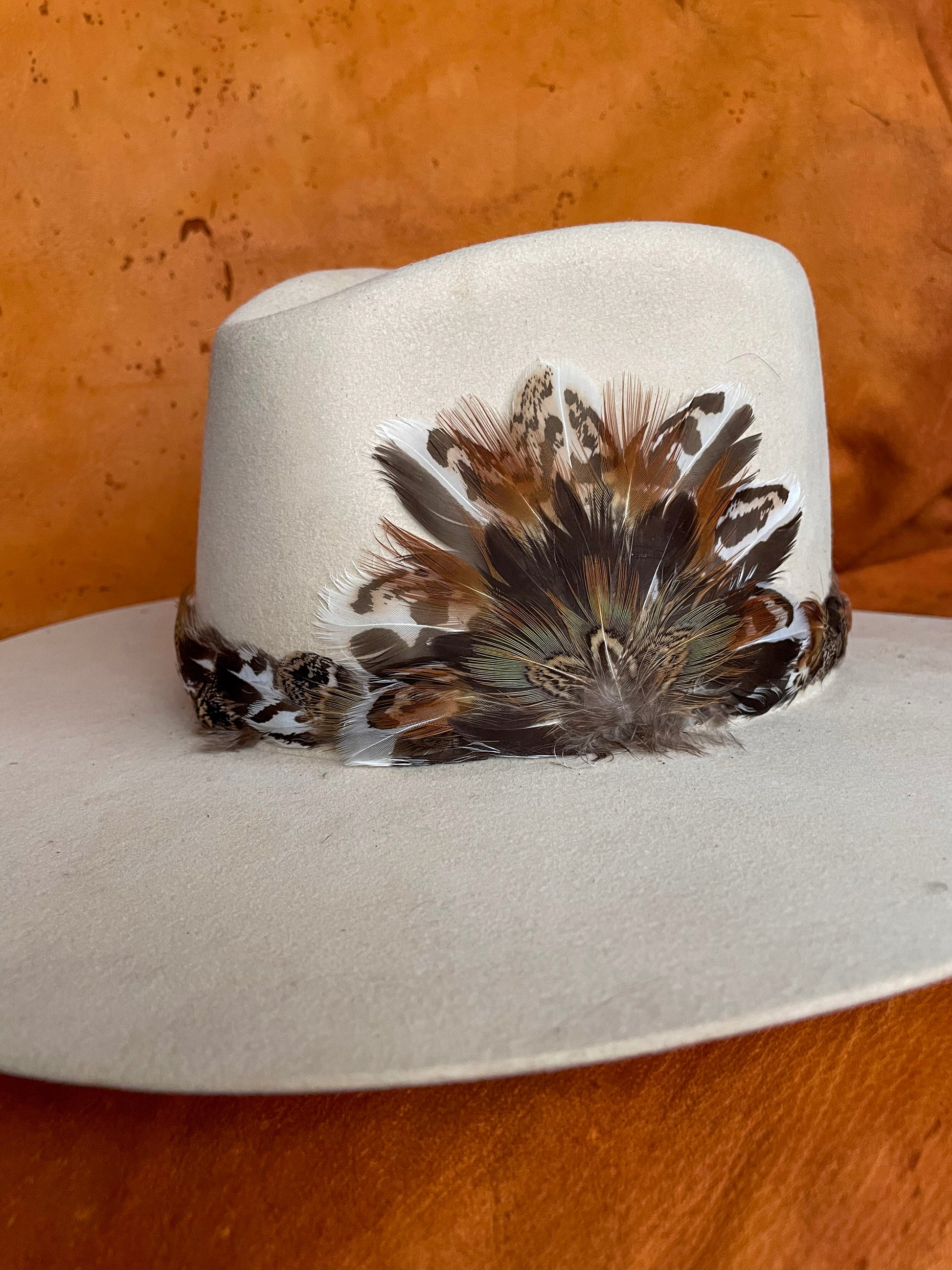Angle view of the high meadow feather hatband and matching removable magnetic medallion. Made with sage grouse feathers and pheasant feathers, shown on a ivory felt hat.  
