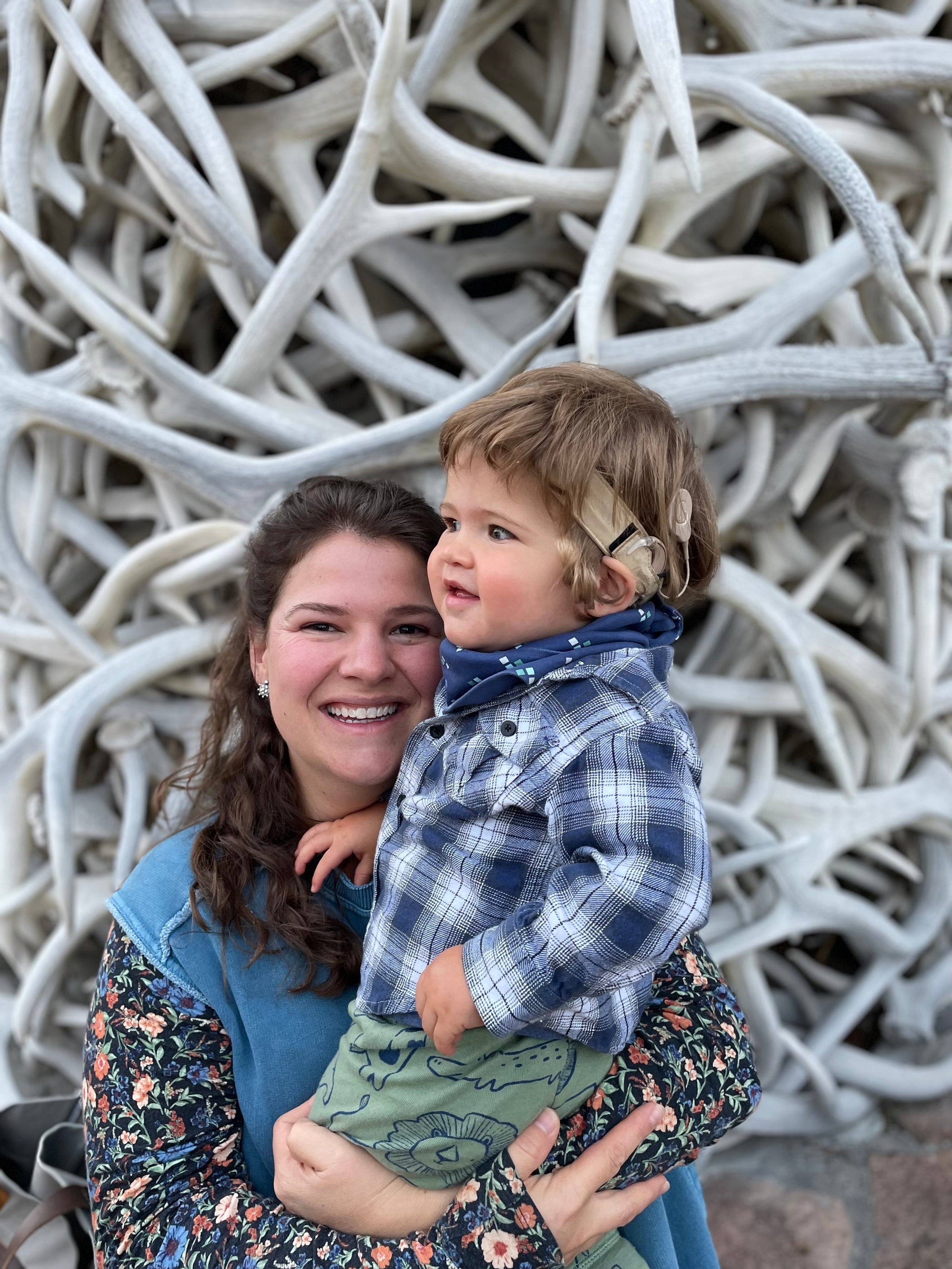 Olivia Poyer smiling at the camera while holding her son Bode in front of Jackson Hole’s iconic elk-antler arches — a joyful snapshot of motherhood and presence in everyday adventure.