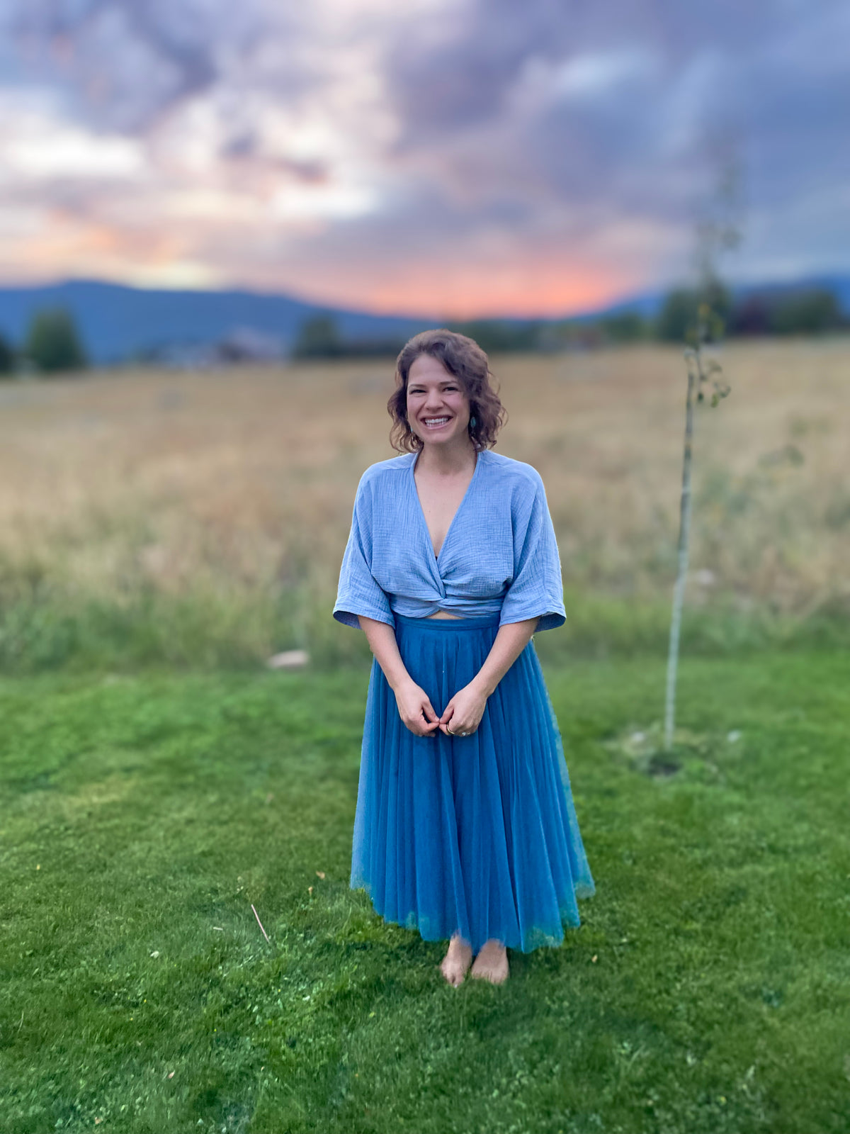 Olivia Poyer smiling in a blue skirt and blouse in a grassy field with mountains and a colorful sunset behind her.