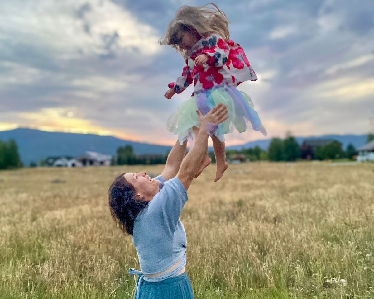Olivia Poyer wearing a blue skirt and tossing her daughter Rosie into the air in a grassy field.