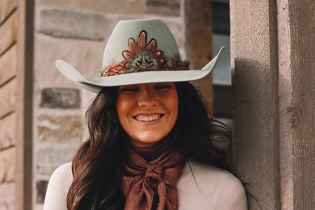 Cowgirl smiling in a Western hat adorned with a handmade feather hatband and medallion by Olivia Poyer.